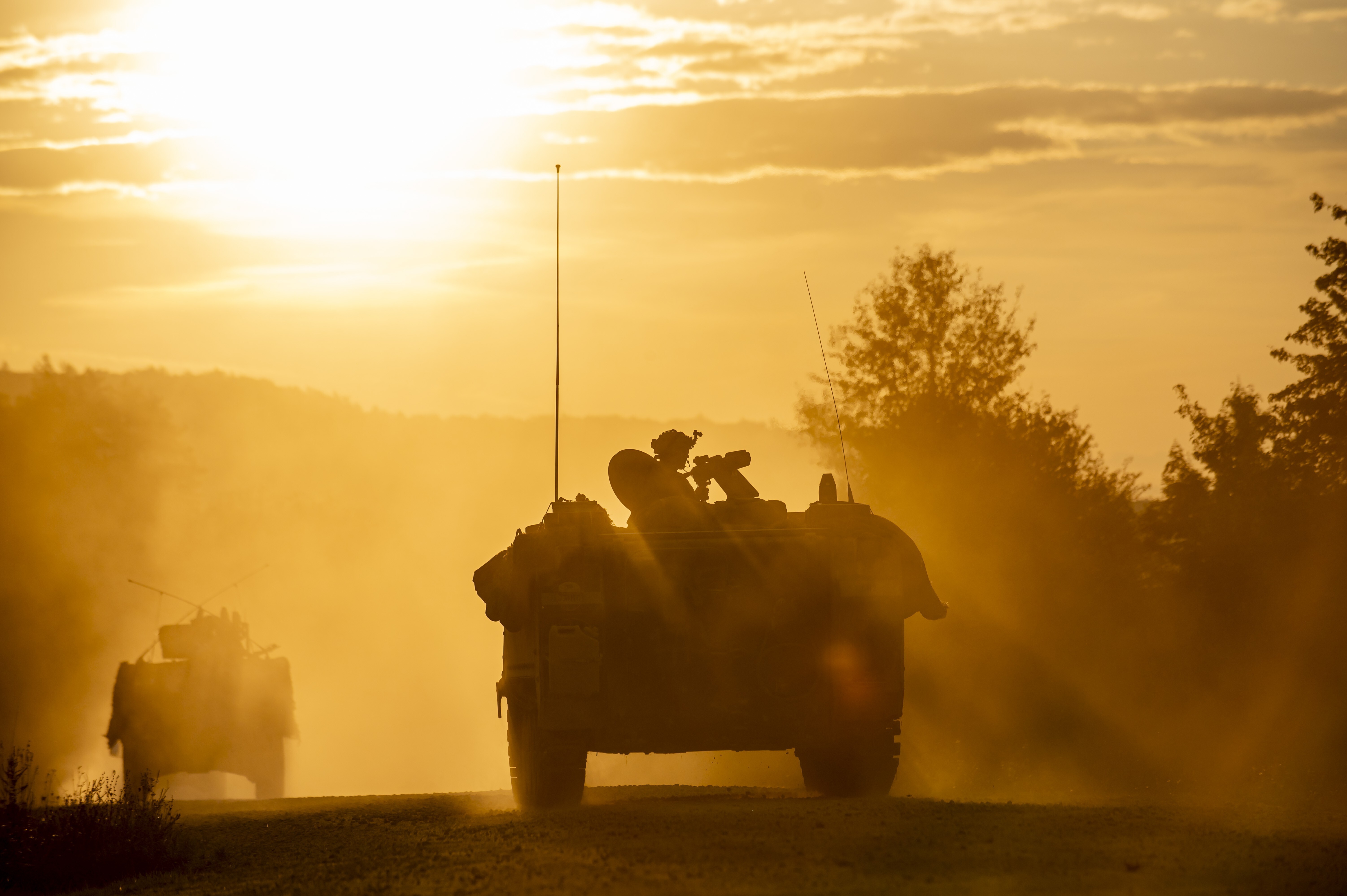 Photograph showing two armored military vehicles driving on a dirt road during sunset, creating a dusty and hazy atmosphere, the type of environmental intrusions that edge AI systems must be protected against. Silhouettes of vehicles and soldiers are visible against a golden sky with scattered clouds and surrounding trees.