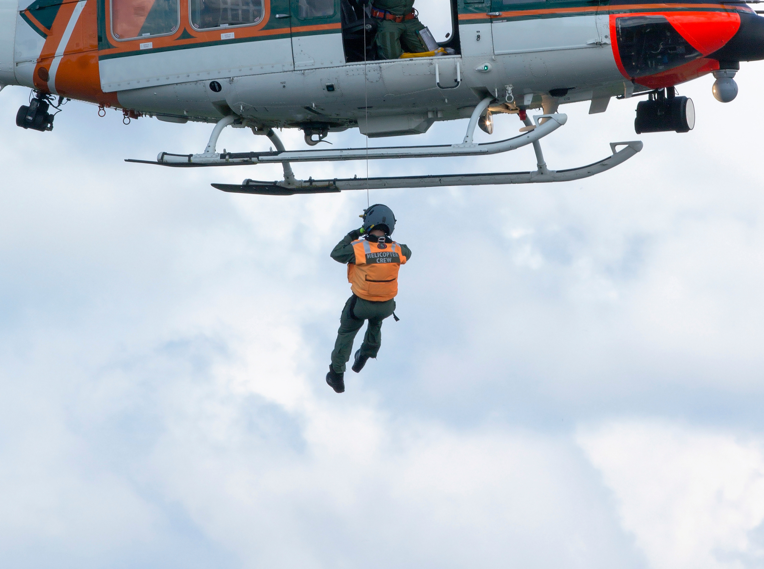 U.S. Coast Guard rescuer wearing a gray uniform and helmet and orange life vest is lowered on a cable from an orange and white helicopter as it hovers.