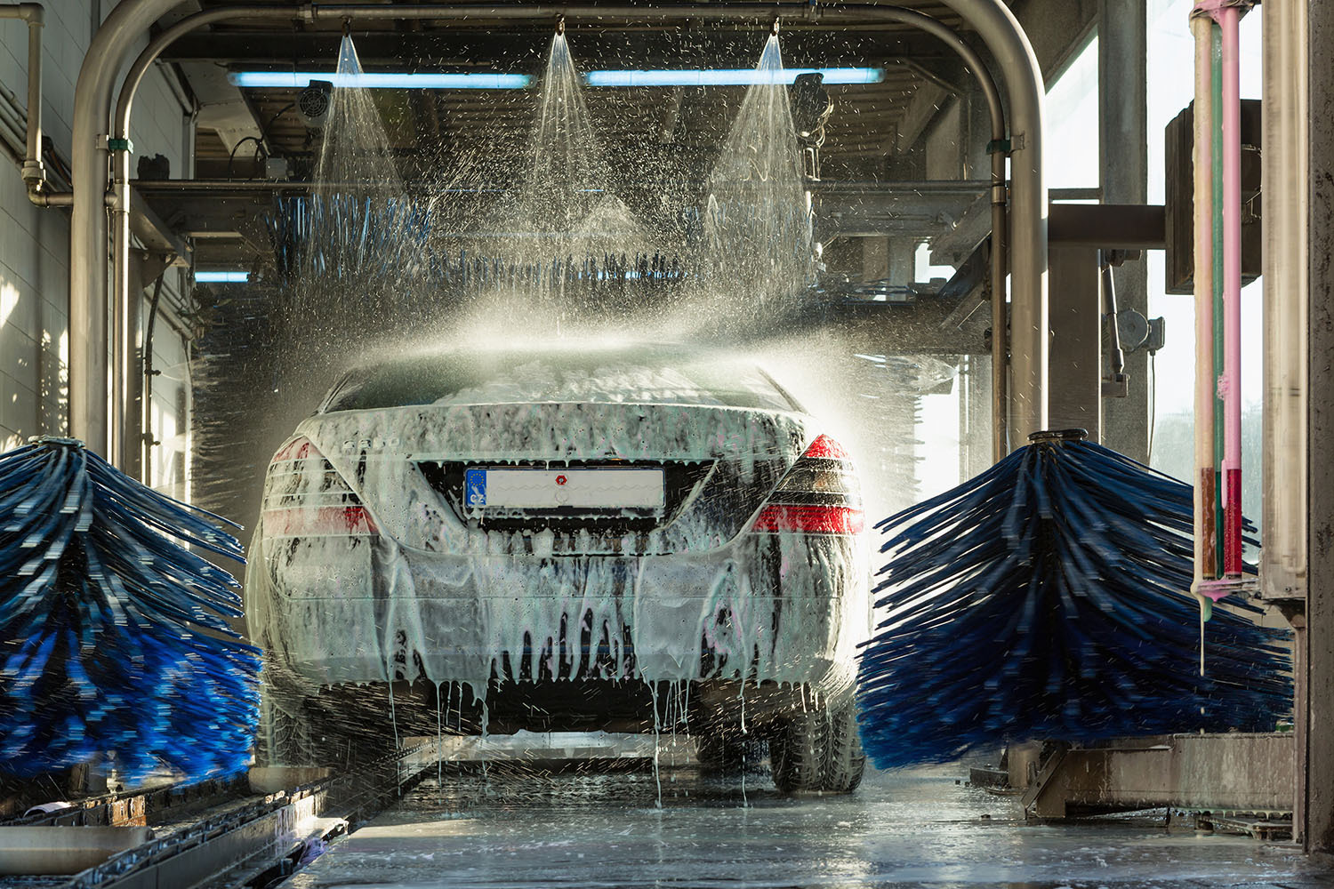Photograph of a car undergoing automated wash inside a car wash station, with soap suds covering rear and water jets spraying from above. Two large rotating blue brushes flank the vehicle, cleaning sides while water and foam drip onto floor.