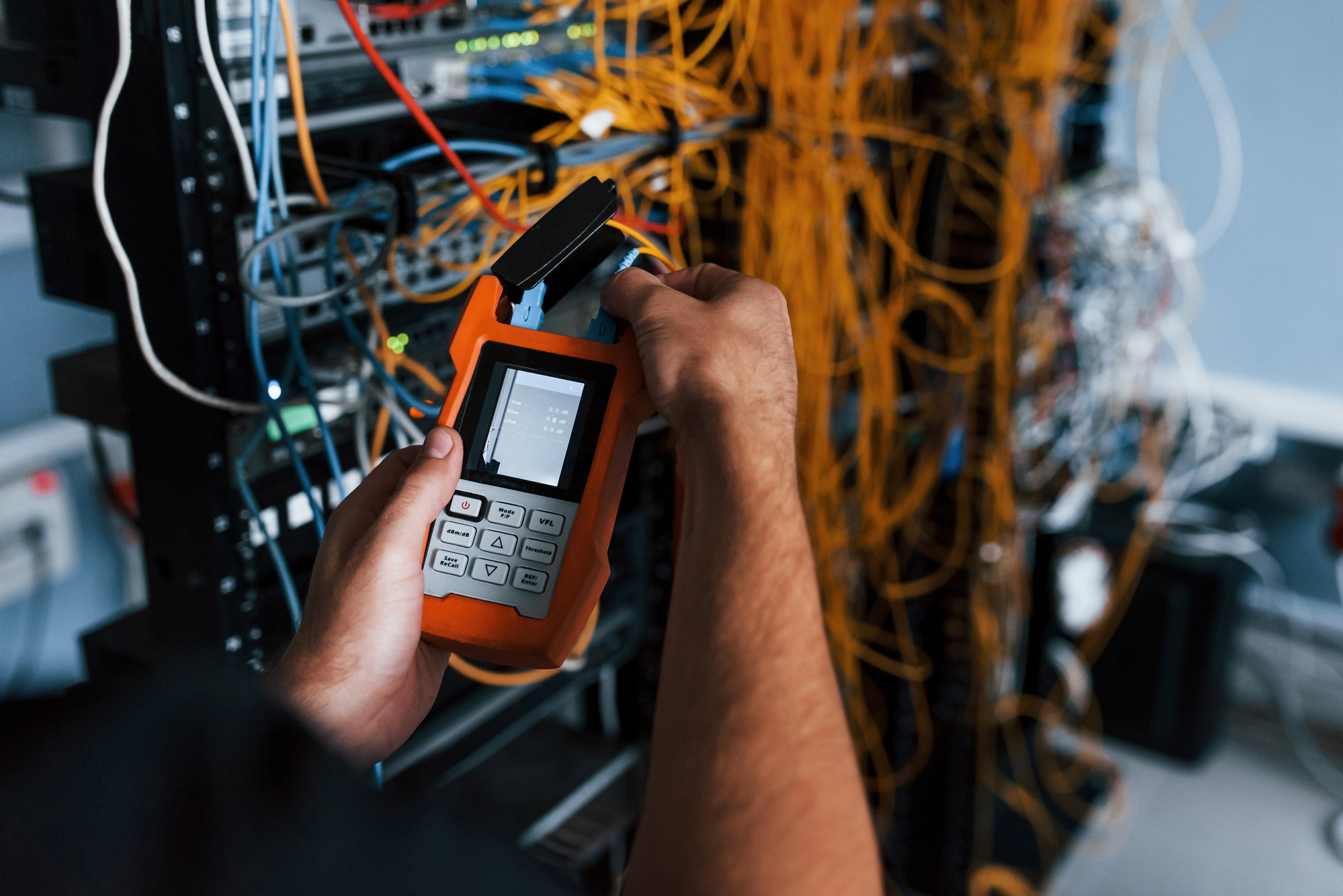 An unorganized bundle of yellow I/O cables on the back of a computer server rack is in the background. Hands holding an orange digital testing device with cables connected to it are in the foreground.