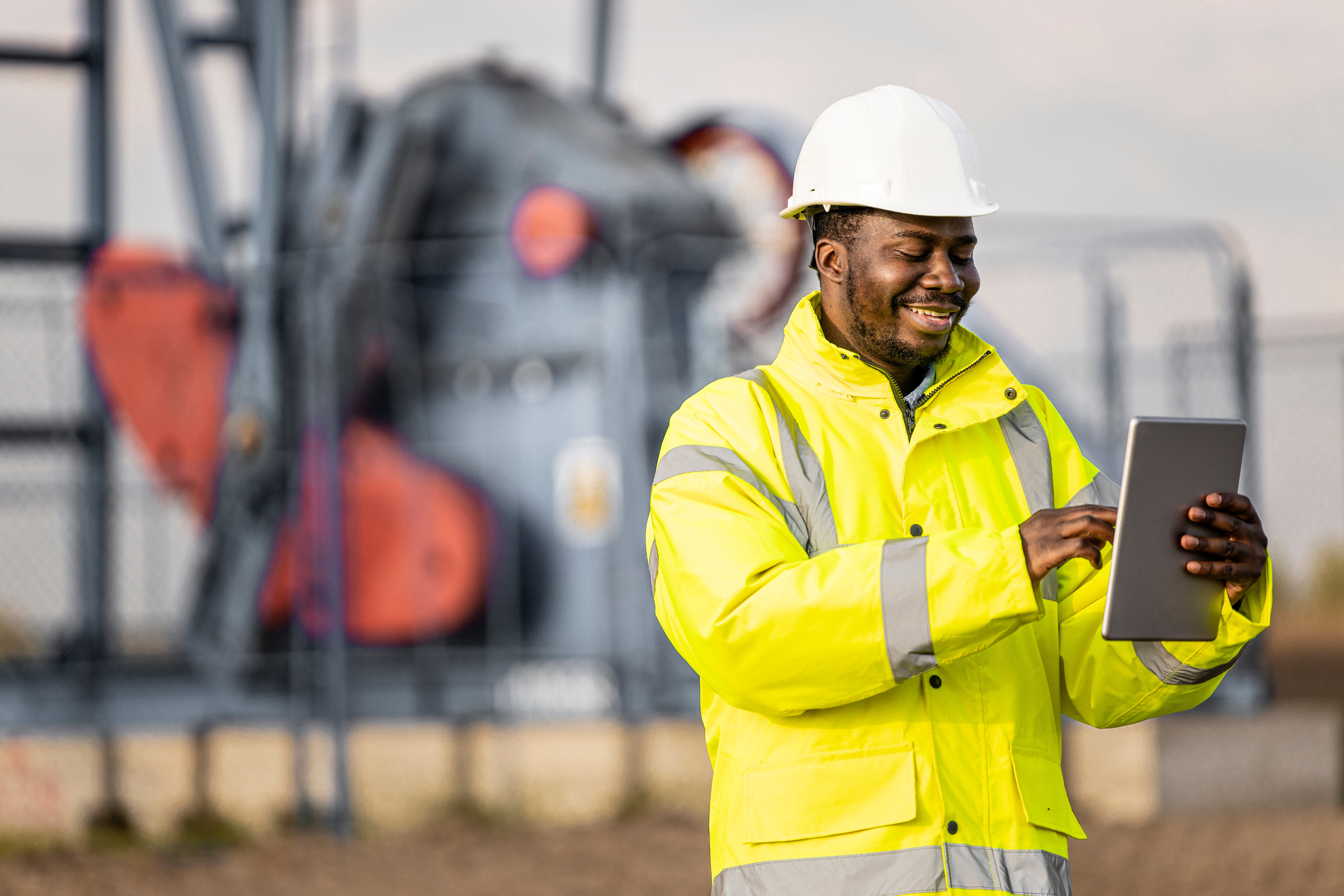 Oil worker in yellow jacket and white hard hat standing and holding a computer while smiling at rig in the field.