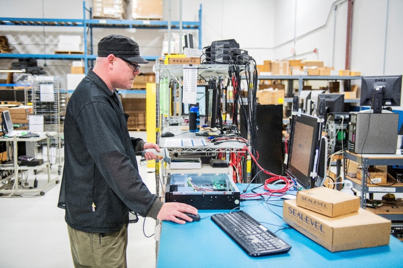 Technician working at a testing station in on a blue table with an open enclosure, sealed component boxes, and organized shelves with various electronic parts in the background.