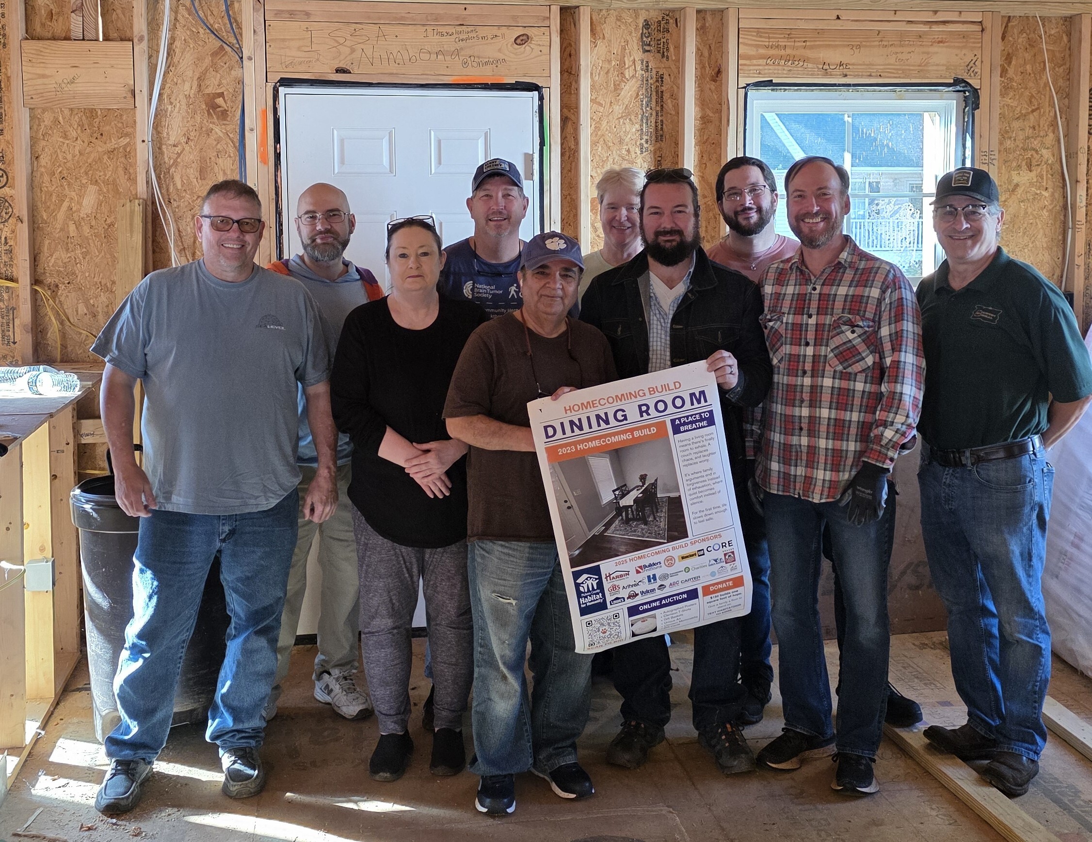 Photograph shows a group of volunteers posing inside an unfinished wood-framed house. One person holds a poster labeled “DINING ROOM,” highlighting a community construction project.