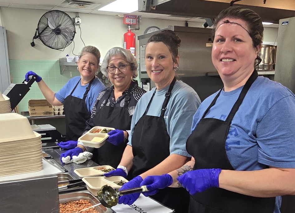 Photograph of four smiling Sealevel Systems employees wearing aprons and purple gloves in a commercial kitchen. Staff assemble and serve meals in compartmented trays beside stacks of plates, prepared food, and kitchen equipment.