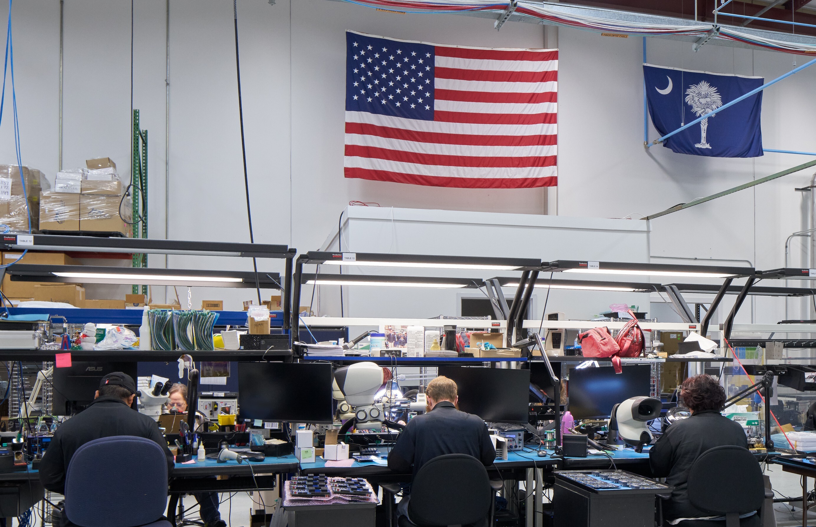 Photograph of a workspace at Sealevel Systems showing three workers seated at desks with multiple monitors, tools, and equipment. American and South Carolina state flags hang on the wall above, indicating location and made in America.